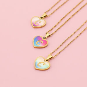 A collection of three initial heart necklaces in different gradient colors laid on a soft pink background.
